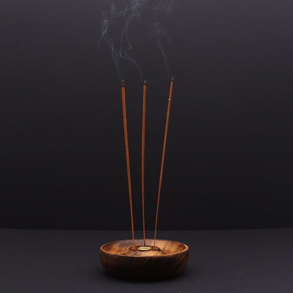 Three burning incense sticks in a wooden bowl holder against a dark background